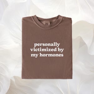 Puede incluir: Una camiseta marr&oacute;n de cuello redondo con el texto blanco personally victimized by my hormones impreso en el centro del pecho. La camiseta tiene un cuello acanalado y una etiqueta de marca visible en el interior del escote.