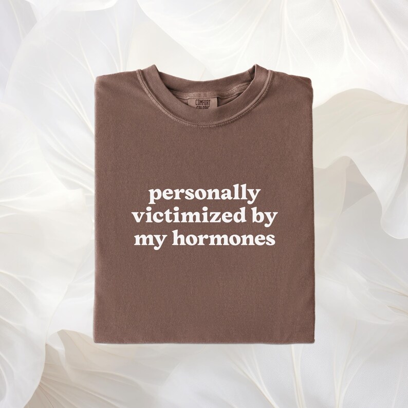 Puede incluir: Una camiseta marr&oacute;n de cuello redondo con el texto blanco personally victimized by my hormones impreso en el centro del pecho. La camiseta tiene un cuello acanalado y una etiqueta de marca visible en el interior del escote.