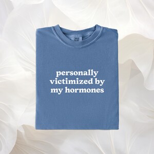 Puede incluir: Una camiseta azul de cuello redondo con el texto blanco personally victimized by my hormones impreso en la parte delantera. La prenda tiene un cuello acanalado y una etiqueta en el escote. La tela tiene un acabado mate y s&oacute;lido en azul.