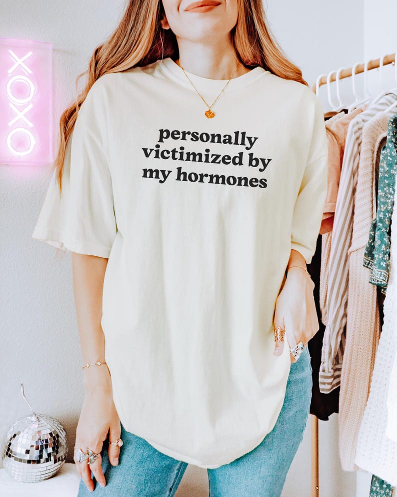 Puede incluir: Una camiseta de manga corta en color blanco roto con el texto negro "personally victimized by my hormones" impreso en una fuente sans-serif sencilla en el pecho. La prenda tiene un corte holgado con un cuello redondo cl&aacute;sico.