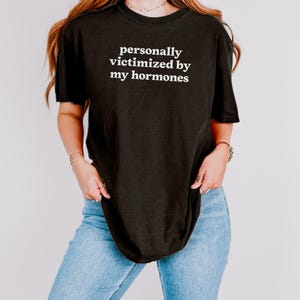 Puede incluir: Una camiseta negra de manga corta con el texto blanco personally victimized by my hormones impreso en el pecho. La prenda tiene cuello redondo y un corte holgado, combinada con unos vaqueros de mezclilla azul claro de cinco bolsillos.