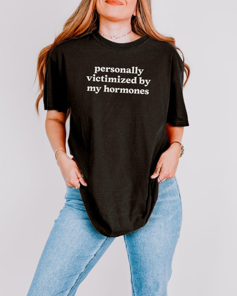 Puede incluir: Una camiseta negra de manga corta con el texto blanco personally victimized by my hormones impreso en el pecho. La prenda tiene cuello redondo y un corte holgado, combinada con unos vaqueros de mezclilla azul claro de cinco bolsillos.