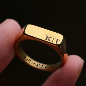 May include: A gold-toned signet ring featuring a flat rectangular top engraved with the letters K|T. The inner band of the ring is engraved with the Roman numeral date II.III.MMXXV. The ring has a polished, smooth metallic finish throughout.