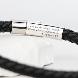 以下が含まれることがあります： 黒い編み込みレザーブレスレット、シルバーの留め金付き。留め金には「私は私を強くしてくださるキリストによって、どんなことでもできるのです。ピリピ4:13」と刻まれています。ブレスレットは白い表面にあります。