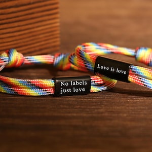 May include: Two rainbow-colored bracelets with black rectangular clasps. One clasp reads "No labels just love," and the other reads "Love is love." The bracelets are made of woven cord and are displayed on a wooden surface.