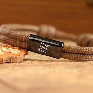May include: A brown rope bracelet with a black rectangular clasp. The clasp features a white graphic of five vertical lines. The bracelet is on a wooden surface.
