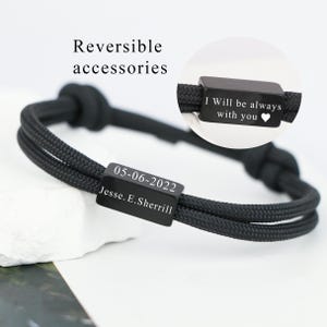 May include: Black bracelet with a woven cord design. The bracelet features a rectangular black metal accent engraved with the date "05-06-2022" and the name "Jesse. E. Sherrill". The text "I Will be always with you" is also engraved.