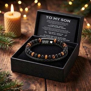 May include: A black gift box containing a beaded bracelet with alternating black and tiger eye stones and a braided black leather band. The clasp features the text SHIELDED BY LOVE and a heart icon. A card inside reads TO MY SON.