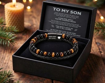 May include: A black gift box containing a bracelet with alternating black and tiger eye beads and a braided black leather band. The metal clasp is engraved with the text SHIELDED BY LOVE and a heart icon. A card inside reads TO MY SON.