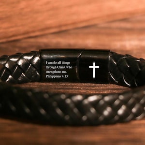 May include: Black braided leather bracelet with a black clasp. The clasp features a white cross and the text "I can do all things through Christ who strengthens me. Philippians 4:13".