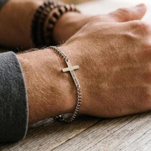 Puede incluir: Una pulsera de cadena de metal plateado con un dije de cruz central. La barra vertical de la cruz está grabada con la fecha 02.03.2026. La pulsera está construida con pequeños eslabones entrelazados y se asegura alrededor de la muñeca.