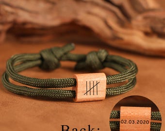 May include: Olive green rope bracelet with a wooden centerpiece. The wooden piece has etched lines, and the back shows the date 02.03.2020. The bracelet is knotted at each end.