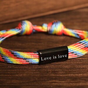 Op de afbeelding: Een regenboogkleurige armband met de tekst "Love is love" op een zwarte rechthoekige sluiting. De armband is gemaakt van gevlochten koord in verschillende kleuren, waaronder rood, oranje, geel, groen, blauw en paars.