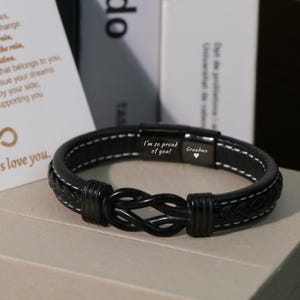 May include: Black leather bracelet with a braided knot design. The bracelet has white stitching and a magnetic clasp engraved with "I'm so proud of you!" and "Grandma". A stylish accessory.
