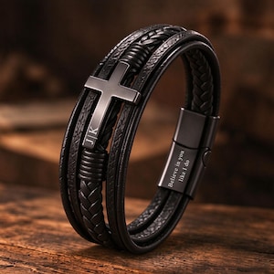 May include: A multi-strand black leather bracelet featuring a central matte black metal cross engraved with the letters J|K. The bracelet includes a black metal magnetic clasp engraved with the text Believe in you like I do, with braided and smooth leather cords.