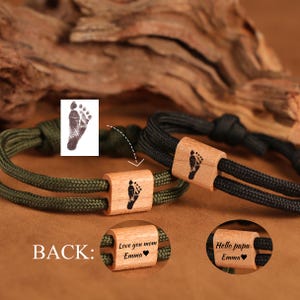 May include: Two bracelets with wooden beads featuring a baby footprint design. One bracelet has an olive green cord, the other a black cord. The wooden beads are engraved with the footprint and personalized messages.
