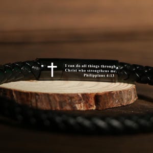 May include: Black braided leather bracelet featuring a silver cross and the inscription "I can do all things through Christ who strengthens me. Philippians 4:13". The bracelet rests on a wooden slice.