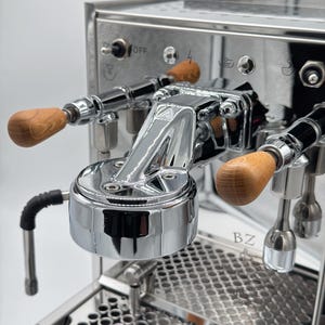 Könnte beinhalten: Nahaufnahme einer Espressomaschine aus Edelstahl mit Holzgriffen. Die Maschine hat eine polierte Chromoberfläche, eine Tropfschale und eine Dampflanze. Der Ein-/Ausschalter ist sichtbar. Die Maschine ist für die Zubereitung von Espresso konzipiert.