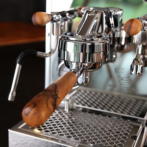 Può includere: Primo piano di una macchina per caffè espresso cromata con manici in legno. Il portafiltro è in primo piano, mostrando una finitura metallica lucida e un manico in legno a motivi. La macchina ha una griglia in acciaio inossidabile.