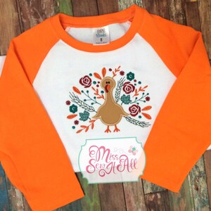 May include: Orange and white long-sleeved shirt with a turkey and floral embroidery. The shirt has a raglan sleeve design. The embroidery features a brown turkey with a red wattle and a floral design with red, green, and white flowers.
