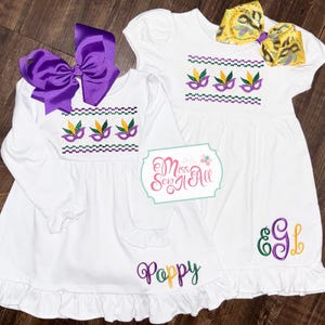May include: Two white dresses with purple, green, and gold embroidery. One dress has the text "Poppy" embroidered on the bottom and the other has the text "E&L" embroidered on the bottom. Both dresses have a row of Mardi Gras masks embroidered on the bodice.
