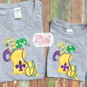 May include: Two heather gray t-shirts featuring an embroidered applique of the state of Louisiana in yellow chevron fabric. The design includes a purple and green jester hat, a purple fleur-de-lis, and green and purple beads. Text reads Miss Sew It All.