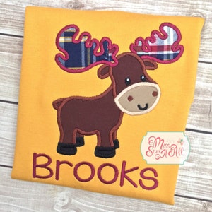 Op de afbeelding: Mosterdgeel T-shirt met een bruin elandontwerp, geruite geweien en de naam "Brooks" geborduurd in bordeauxrood. De eland heeft een bruine snuit en zwarte hoeven. Klein logo in de hoek.