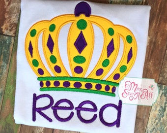 May include: White t-shirt with a yellow and green crown embroidered on it. The crown has purple accents and the name "Reed" is embroidered below it in purple.
