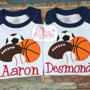 May include: Two white raglan shirts with navy blue sleeves, each featuring a soccer ball, basketball, football, and baseball applique. One shirt says "Aaron" in red, the other says "Desmond".