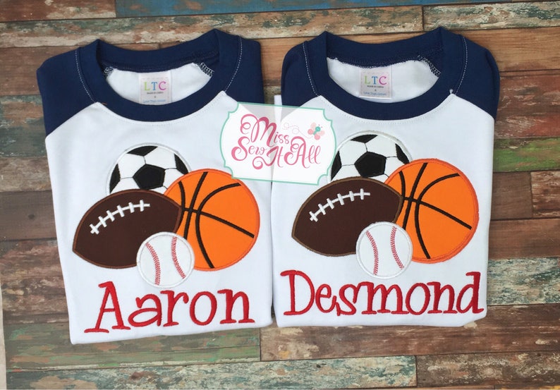 May include: Two white raglan shirts with navy blue sleeves, each featuring a soccer ball, basketball, football, and baseball applique. One shirt says "Aaron" in red, the other says "Desmond".