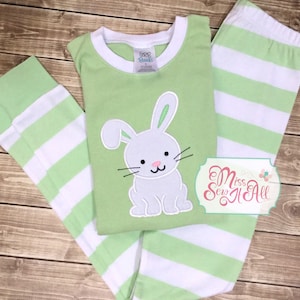 May include: Green and white striped pajamas with a white bunny applique on the shirt. The bunny has green ears and a pink nose. The shirt has the text "Crib Friends" on the tag.