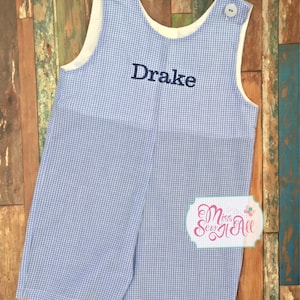 May include: A blue and white gingham overall with the name "Drake" embroidered on the front. The overall has a white lining and two buttons on the straps.