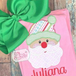 May include: Pink Christmas outfit with a Santa Claus applique and the name "Juliana" embroidered in red. A large green bow is attached. The outfit is likely for a child.