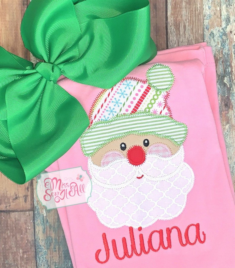 May include: Pink Christmas outfit with a Santa Claus applique and the name "Juliana" embroidered in red. A large green bow is attached. The outfit is likely for a child.
