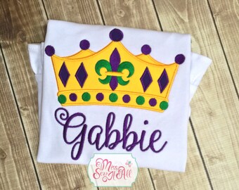 May include: White t-shirt with a purple, yellow, and green embroidered crown and the name "Gabbie" in purple cursive lettering. The crown features a fleur-de-lis in the center.