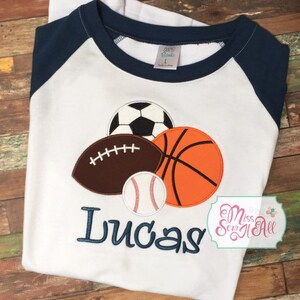 May include: White baseball-style shirt with navy blue sleeves. The shirt features appliqued sports balls: a soccer ball, football, basketball, and baseball. The name "Lucas" is embroidered in navy blue thread.