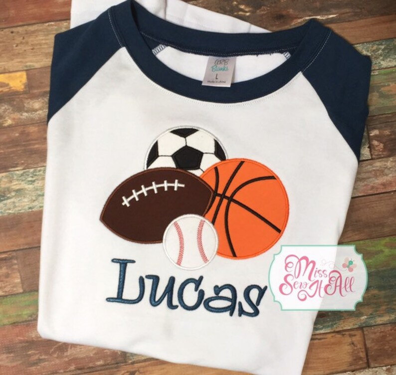 May include: White baseball-style shirt with navy blue sleeves. The shirt features appliqued sports balls: a soccer ball, football, basketball, and baseball. The name "Lucas" is embroidered in navy blue thread.