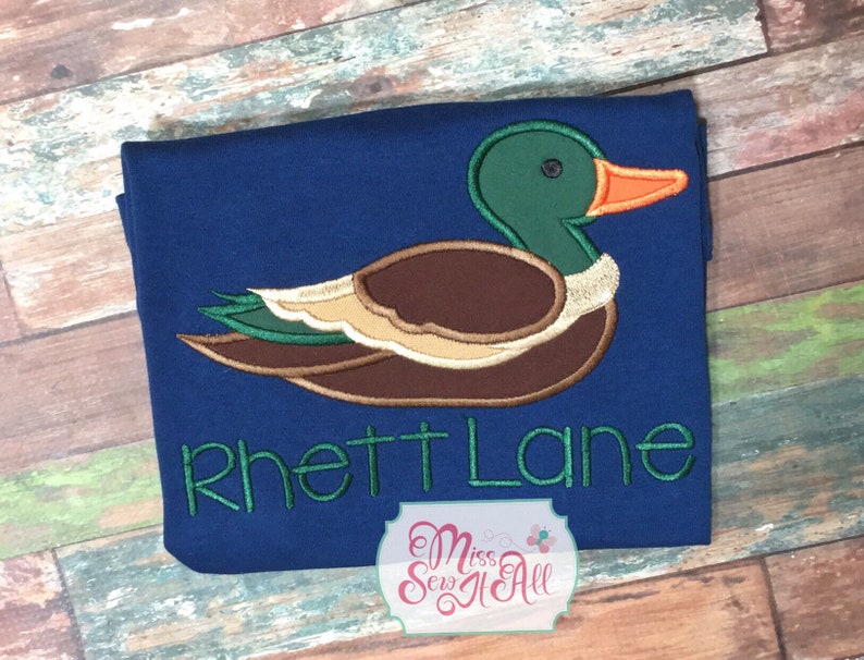 以下が含まれることがあります： 紺色の生地に刺繍されたアヒルのデザイン。アヒルは緑色の頭、茶色の体、オレンジ色のくちばしをしています。「Rhett Lane」という名前が緑色で刺繍されています。小さなロゴが右下にあります。