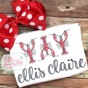 May include: White t-shirt with three embroidered red lobsters and the text "ellis claire" in gray cursive lettering.