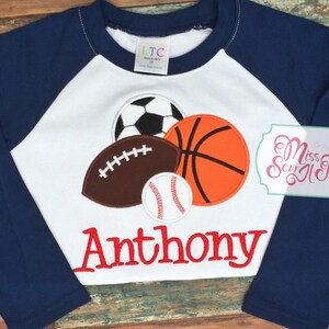 May include: A navy blue and white raglan-style long-sleeve shirt featuring appliqued sports balls: a soccer ball, football, basketball, and baseball. The name "Anthony" is embroidered in red script below the sports graphics.