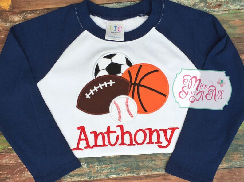 May include: A navy blue and white raglan-style long-sleeve shirt featuring appliqued sports balls: a soccer ball, football, basketball, and baseball. The name "Anthony" is embroidered in red script below the sports graphics.