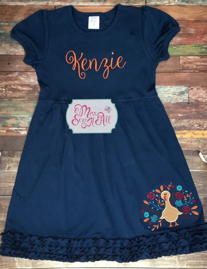 May include: A navy blue dress with a ruffled hem. The dress has an orange embroidered turkey on the bottom right side and the name "Kenzie" embroidered on the front in orange. The dress also has a white tag with the text "Miss Sew It All" on the front.
