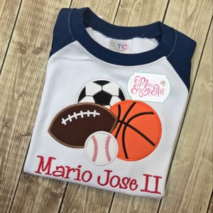 May include: White and navy blue raglan style t-shirt featuring appliqued sports balls: a soccer ball, football, basketball, and baseball. The name "Mario Jose II" is embroidered in red script across the bottom. The shirt has a round neck.