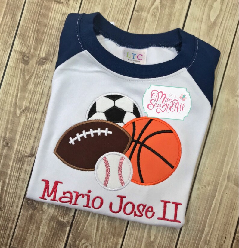 May include: White and navy blue raglan style t-shirt featuring appliqued sports balls: a soccer ball, football, basketball, and baseball. The name "Mario Jose II" is embroidered in red script across the bottom. The shirt has a round neck.