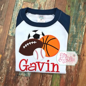 May include: White and navy blue raglan style shirt with sports ball appliqu&eacute;s including a soccer ball, football, basketball, and baseball. The name "Gavin" is embroidered in red. The shirt has a tag that says "5T".
