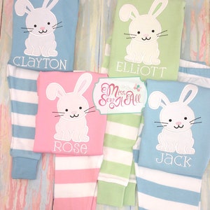 May include: Four pairs of striped pajamas with a white bunny applique on the top. The pajamas are in blue, green, pink, and light blue. The names Clayton, Elliott, Rose, and Jack are embroidered on the tops.