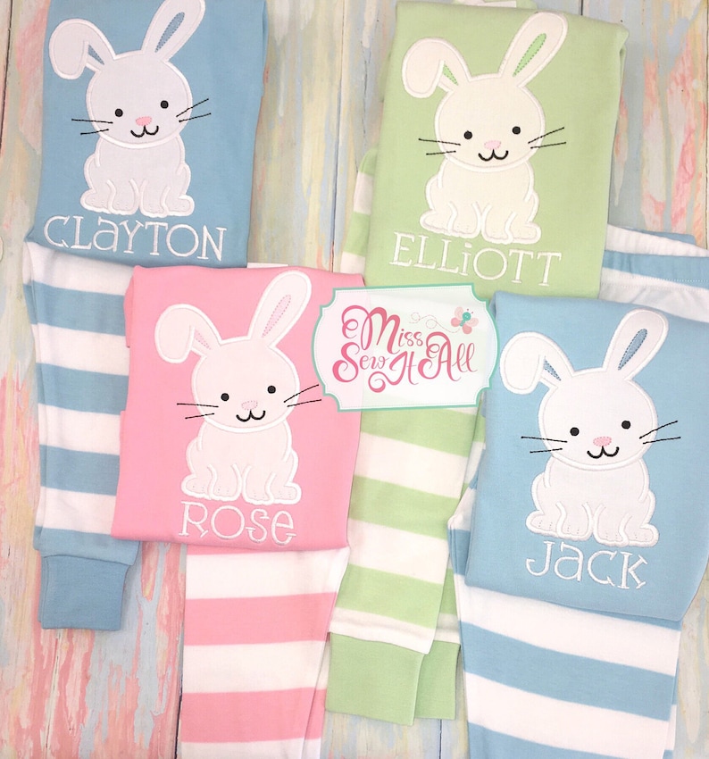 May include: Four pairs of striped pajamas with a white bunny applique on the top. The pajamas are in blue, green, pink, and light blue. The names Clayton, Elliott, Rose, and Jack are embroidered on the tops.