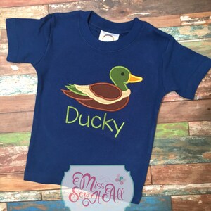 以下が含まれることがあります： 刺繍されたアヒルのデザインが施された、濃いネイビーブルーの半袖Tシャツ。アヒルは緑色の頭、茶色の体、黄色のくちばしをしています。「Ducky」の文字が緑色で刺繍されています。子供服です。