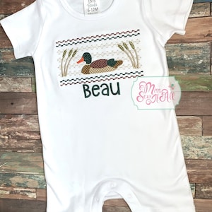 May include: White baby romper with a duck design. The design features a duck, cattails, and the name "Beau" in green. The romper has short sleeves and snaps at the bottom. The tag reads "6-12M".