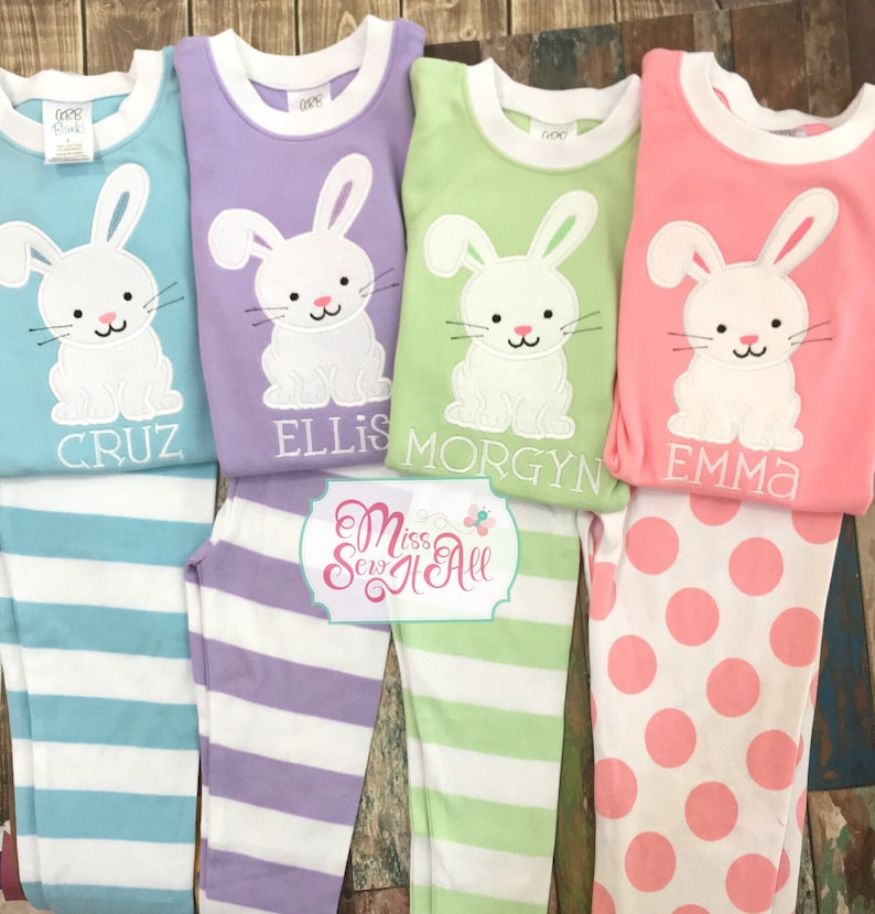 May include: Four pairs of children's pajamas with bunny appliques and personalized names. The pajamas are in pink, purple, green, and blue with matching striped or polka dot pants.